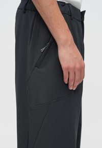 Tumši pelēkas bikses no gludas, elastīgas materiāla. Tās ir ar slīpu priekšējo kabatu ar rāvējslēdzēju un vaļēju piegriezumu jostasvietā.