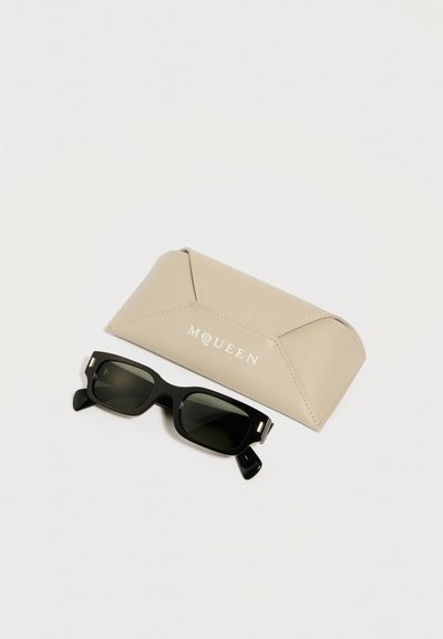 Μαύρα ορθογώνια γυαλιά ηλίου με σκοτεινούς φακούς δίπλα σε μπεζ δερμάτινη θήκη με την επιγραφή "McQueen" σε λευκά γράμματα.