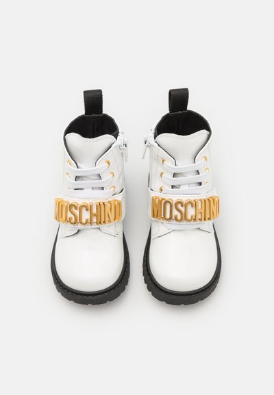 Valged lakknahast pahkluu saapad mustade talladega, millel on kuldne "MOSCHINO" logo rihm, valged paelad ja külgkattega tõmblukk.