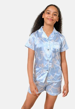 Ung pige med krøllet hår iført lyseblåt satin kortærmet knapbluse og matchende shorts med hvide abstrakte mønstre.