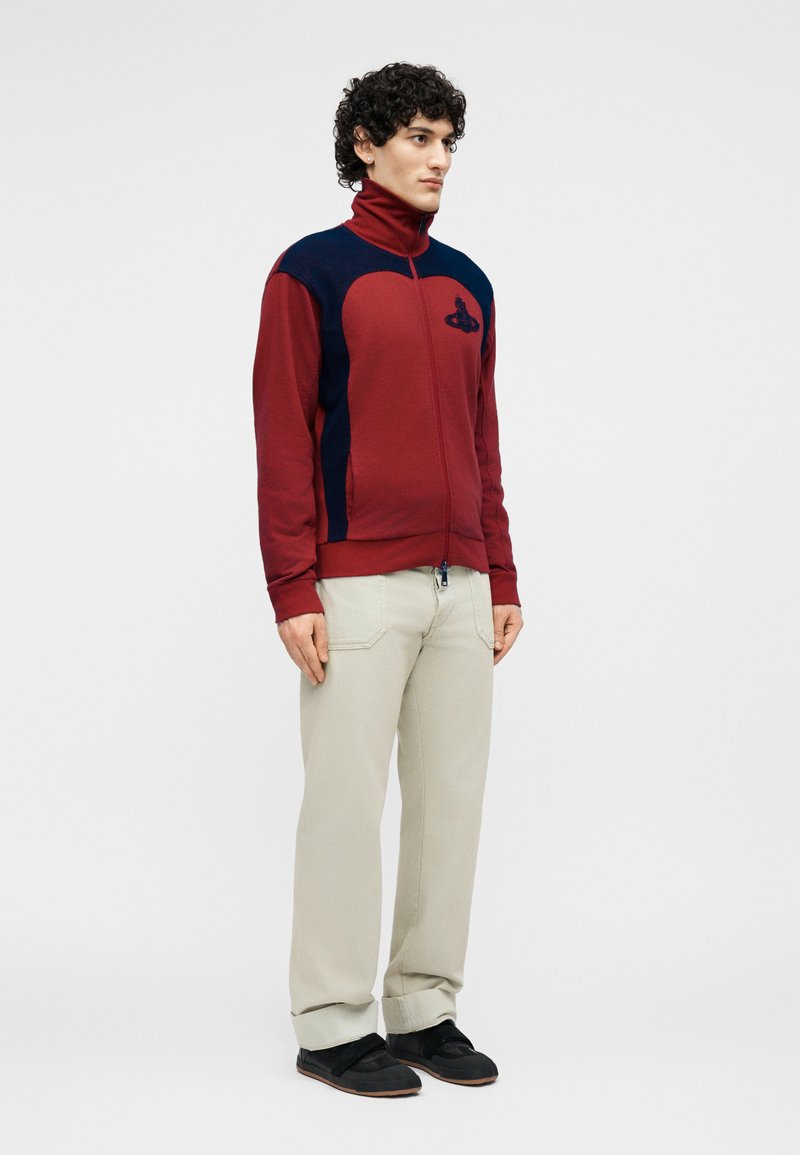 Man standing wearing a red and navy zipped jacket, beige pants, and black shoes against a plain light background.