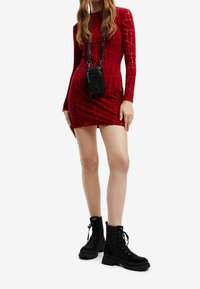 Rotes Samtkleid mit langen Ärmeln und strukturiertem Muster, tailliertes Design. Accessoire: kleine schwarze strukturierte Tasche. Schuhe: schwarze Schnürkampfstiefel.