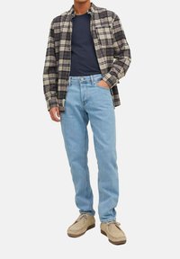 Rutet flannel skjorte i beige og sort over en marineblå t-shirt, parret med lyseblå jeans og beige ruskindsko. Klassisk pasform og afslappet stil.