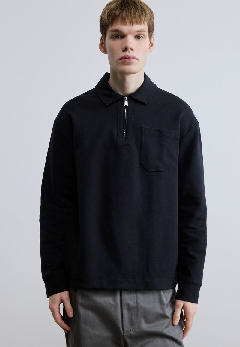 Μαύρο πόλο με μακριά μανίκια και φερμουάρ στο γιακά, με τσέπη μπροστά. Λείο ύφασμα και άνετη εφαρμογή.