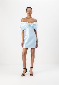 Abito in satin azzurro chiaro con scollatura a spalle scoperte e grande dettaglio a fiocco al petto; design aderente, lunghezza corta, texture liscia, abbinato a sandali neri con tacco.