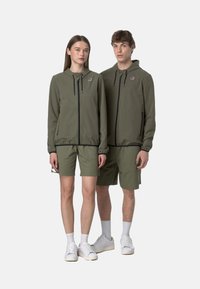 Giacca a zip verde oliva con cappuccio e pantaloni corti abbinati, caratterizzata da una zip nera a contrasto e tasche laterali, indossata con sneakers bianche.