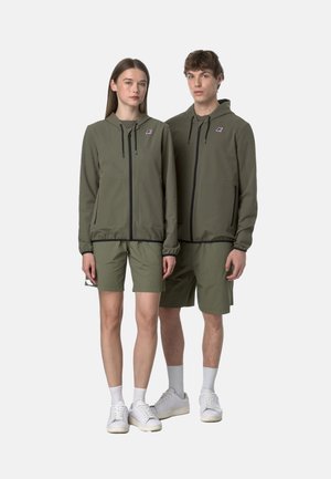 Giacca a zip verde oliva con cappuccio e pantaloni corti abbinati, caratterizzata da una zip nera a contrasto e tasche laterali, indossata con sneakers bianche.