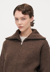Brun, zip-up jacka med hög krage, tillverkad av strukturerat tyg. Har en silverfärgad dragkedja och en avslappnad passform.