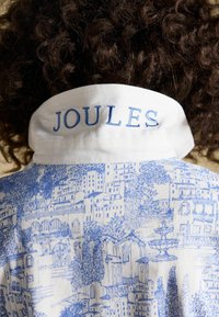 Persoană cu păr creț purtând o cămașă cu guler alb, cu imprimeu albastru cu peisaj urban și „JOULES” brodat cu albastru pe partea din spate a gulerului.