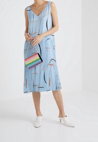 Abito plissettato azzurro chiaro con moderni motivi astratti neri e marroni, scollatura a V. La modella tiene una borsa metallizzata color arcobaleno. Mules bianche.