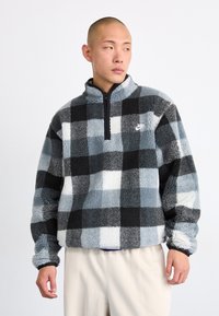 Polár pulóver fekete, szürke és fehér kockás mintával; félig cipzáras gallér; Nike logó a bal mellen; texturált, puha anyag; laza szabás.