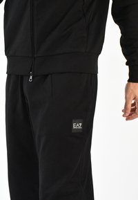 Chaqueta negra con cremallera y pantalones a juego, hechos de material suave; presentan detalles de costura y un parche con el logotipo en los pantalones.