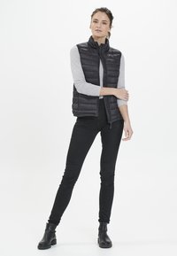 Chaleco acolchado negro con cuello alto, frente con cremallera y material ligero, combinado con una camiseta de manga larga gris y jeans ajustados negros.