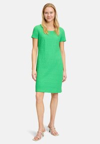 Grünes kurzärmliges Kleid mit strukturiertem Strickdesign, geradem Schnitt, knieumspielender Länge und rundem Ausschnitt. Kombiniert mit beigen Absatzsandalen.
