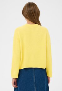 Pull jaune surdimensionné avec une finition douce et texturée, des poignets côtelés et un ourlet court, associé à une jupe en denim.