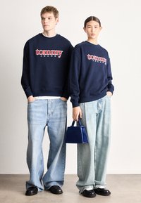 Námornícke mikiny s logom "tommy JEANS" nosené cez svetlomodré džíny s širokými nohavicami. Modelka drží malú modrú kabelku; čierne topánky dotvárajú outfit.