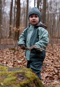 Vattentät grön och grå jacka och byxor, med en brun huva, båda på en barn som håller i en pinne, omgivet av en skog och fallna löv.