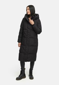 Sort polstret lang frakke med hætte, præget af et quiltet mønster, lodrette syninger og trykknaplukning. Sammen med sorte ankelstøvler.