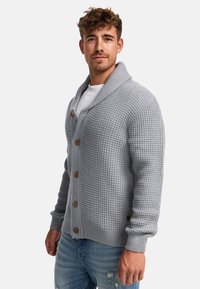 Grau gestrickte Strickjacke mit einem Schalkragen, großen vorderen Knöpfen und gerippten Bündchen. Gewebte Textur, über einem weißen T-Shirt getragen, kombiniert mit blauen Jeans.
