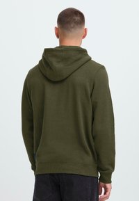 Joven con pelo corto, vestido con una sudadera verde oliva y pantalones oscuros, de pie de espaldas contra un fondo claro y liso.