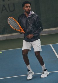 Svart texturerad jacka över en lila skjorta med krage, vita shorts och vita sneakers med svarta och gröna detaljer. Håller en orange tennisracket.