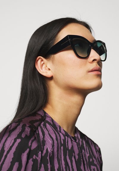 Sorte cat-eye solbriller med tonede linser, tykke rammer og vinkelagtigt design, båret af en model iført en top med lilla og sorte mønstre.