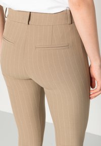 Pantaloni beige su misura con sottili righe bianche, dotati di due tasche posteriori e di un tessuto liscio e texturizzato. Design a vita alta.