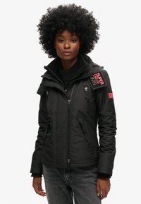 Sort zip-jakke med høj krave, frontlynlås, flere lommer, stribemønster og broderet logo på skulderen.