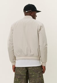 Lys beige bomberjakke med ribbet kant, glat tekstur og en sort kasket. Båret over et hvidt mesh-plagg lagd med camouflaged bukser.