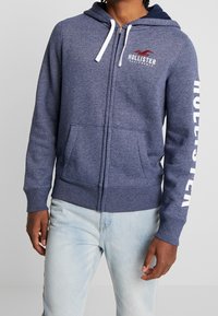 Modell iförd en mörk marinblå zip-up hoodie med vita dragsnören och "Hollister"-logotyp på bröstet och ärmen, kombinerat med ljusblå jeans.