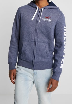 Modello che indossa una felpa con zip di un blu scuro con lacci bianchi e il logo "Hollister" sul petto e sulla manica, abbinata a jeans azzurri chiari.