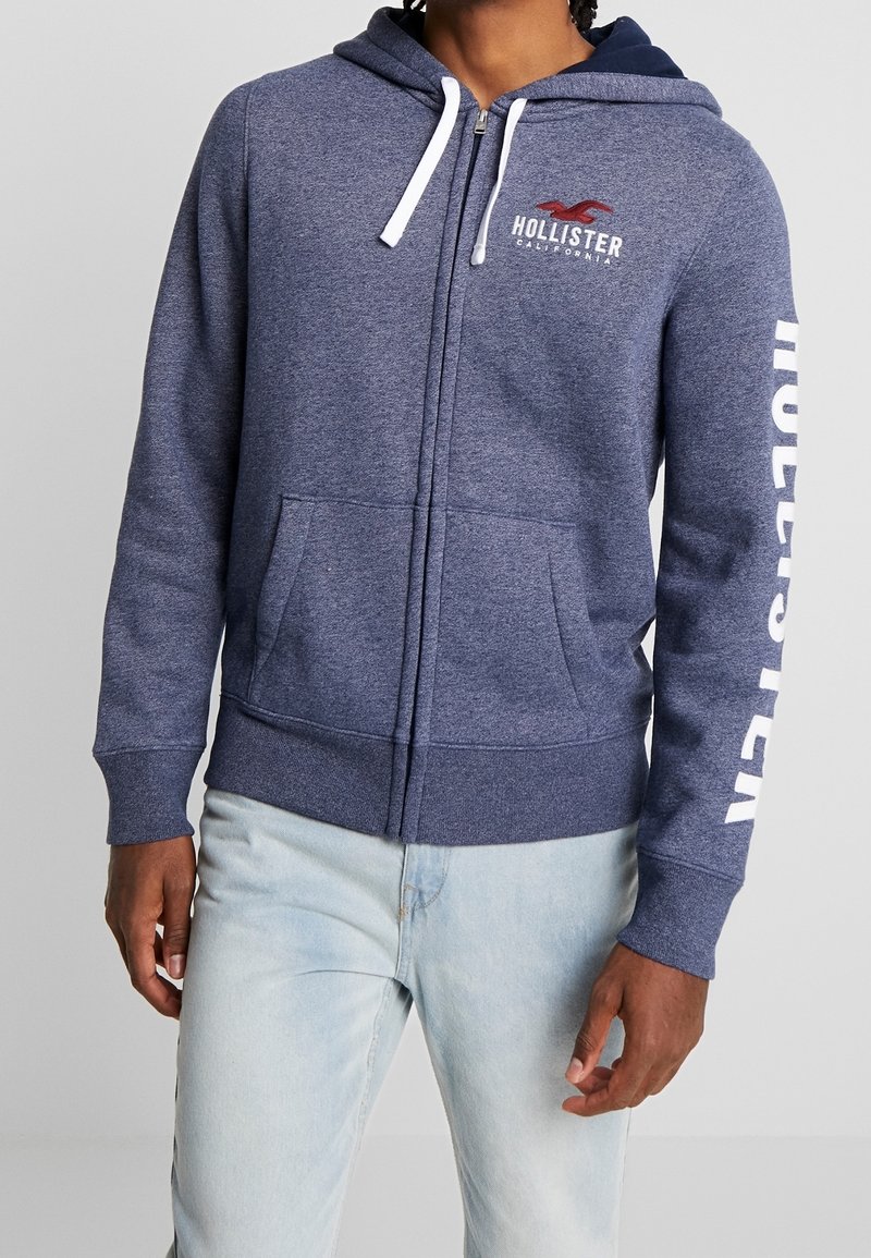 Modell iförd en mörk marinblå zip-up hoodie med vita dragsnören och "Hollister"-logotyp på bröstet och ärmen, kombinerat med ljusblå jeans.