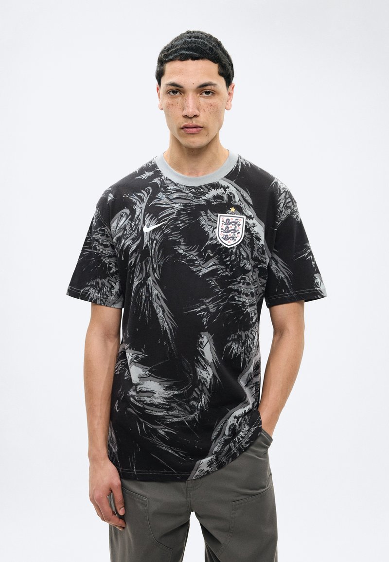 Νέος άνδρας με κοντά σκούρα μαλλιά, ντυμένος με μαύρη και γκρι μπλούζα ποδοσφαίρου της Αγγλίας με το έμβλημα των τριών λιονταριών και γκρι παντελόνι.