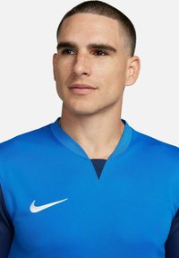 Le maillot de sport bleu présente un design à col en V avec des manches navy contrastantes. Logo Nike blanc sur la poitrine. Tissu lisse et léger.