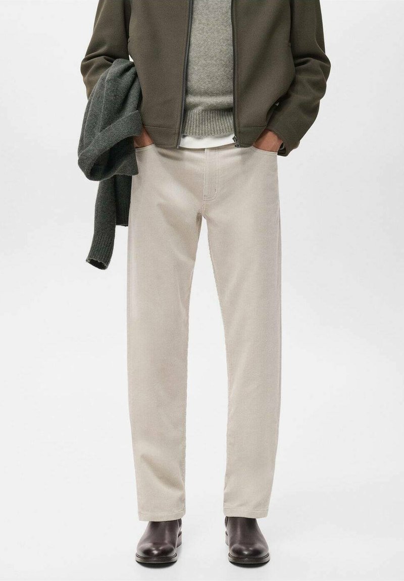 Pantaloni in corduroy beige con vestibilità dritta, caratterizzati da una sottile texture a costine. Abbinati a un maglione grigio e a una giacca verde scuro, indossati con stivali marroni.