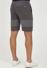 Pantalones cortos deportivos grises con paneles en gris más oscuro, que cuentan con un bolsillo trasero y una tela suave y texturizada. Las zapatillas blancas complementan el conjunto.