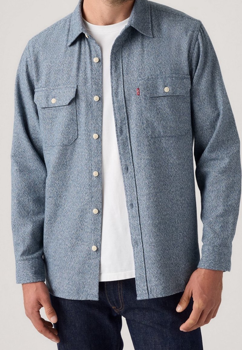 Homme portant une chemise à boutons bleu clair par-dessus un t-shirt blanc et un jean bleu foncé, debout les bras détendus le long du corps.