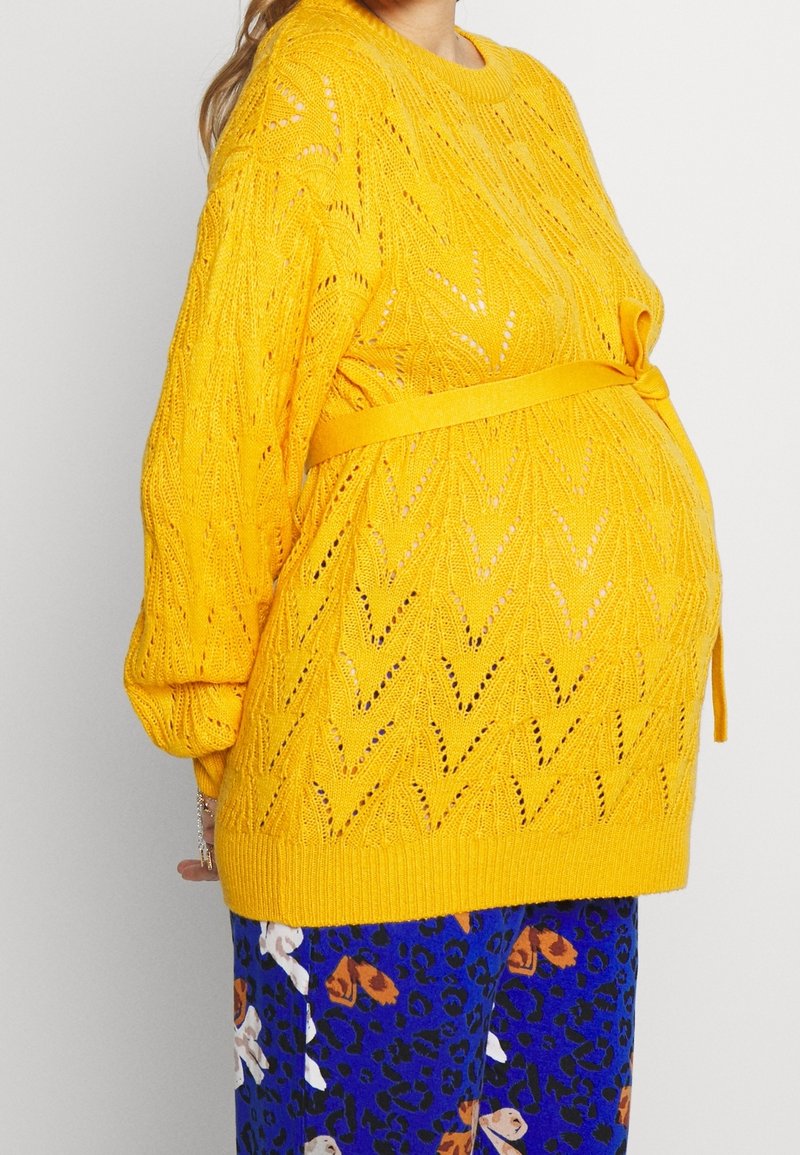 Personne enceinte portant un pull en tricot jaune vif avec une ceinture nouée et un pantalon bleu à motifs, debout devant un fond uni.