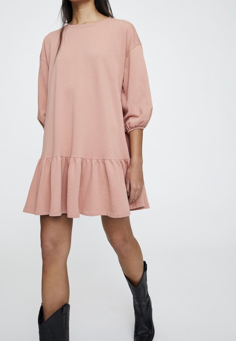 Rosa Baumwollkleid mit dreiviertellangen Puffärmeln, gerafftem Saum und lockerer Silhouette, kombiniert mit schwarzen Cowboy-Stiefeln.