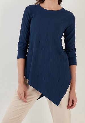 Femme portant un haut bleu marine texturé à manches longues avec un ourlet asymétrique, associé à un pantalon beige clair sur fond uni.