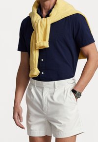 Camicia azzurra a maniche corte, pantaloni bianchi con pieghe e un maglione giallo a trecce appoggiato sulle spalle. Orologio nero con quadrante rotondo.