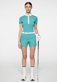 Turkos golfpolo med vita accenter och krage, paired with matching shorts. Tillbehör inkluderar ett vitt bälte och en handske, som håller en golfklubba.