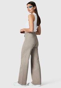Donna con capelli lunghi e scuri che indossa un trucco occhi geometrico blu, top bianco senza maniche, pantaloni beige a gamba larga e scarpe bianche, in piedi di lato.