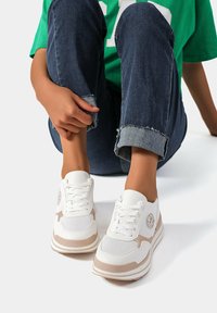 Sneaker sportive bianche con accenti beige, pannelli in rete e design con lacci. Punta rotonda e suola ammortizzata, posizionate su jeans scuri.