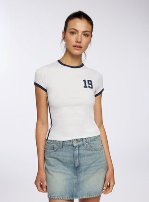 Giovane donna con una t-shirt bianca aderente con bordino navy e stampa "19", abbinata a una minigonna di jeans azzurro chiaro, in piedi davanti a uno sfondo semplice.