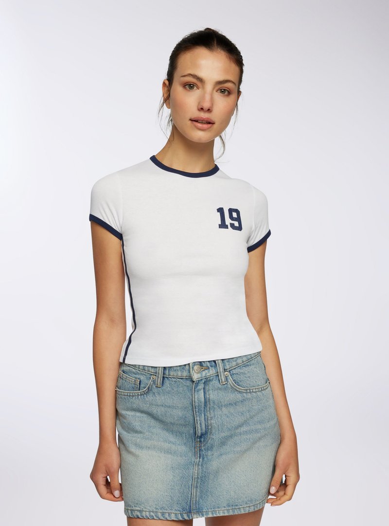 Giovane donna con una t-shirt bianca aderente con bordino navy e stampa "19", abbinata a una minigonna di jeans azzurro chiaro, in piedi davanti a uno sfondo semplice.
