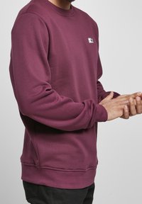 Hombre vestido con un sudadera de cuello redondo, de manga larga y color burdeos, con un pequeño logo blanco en el pecho, ajustándose la manga izquierda.
