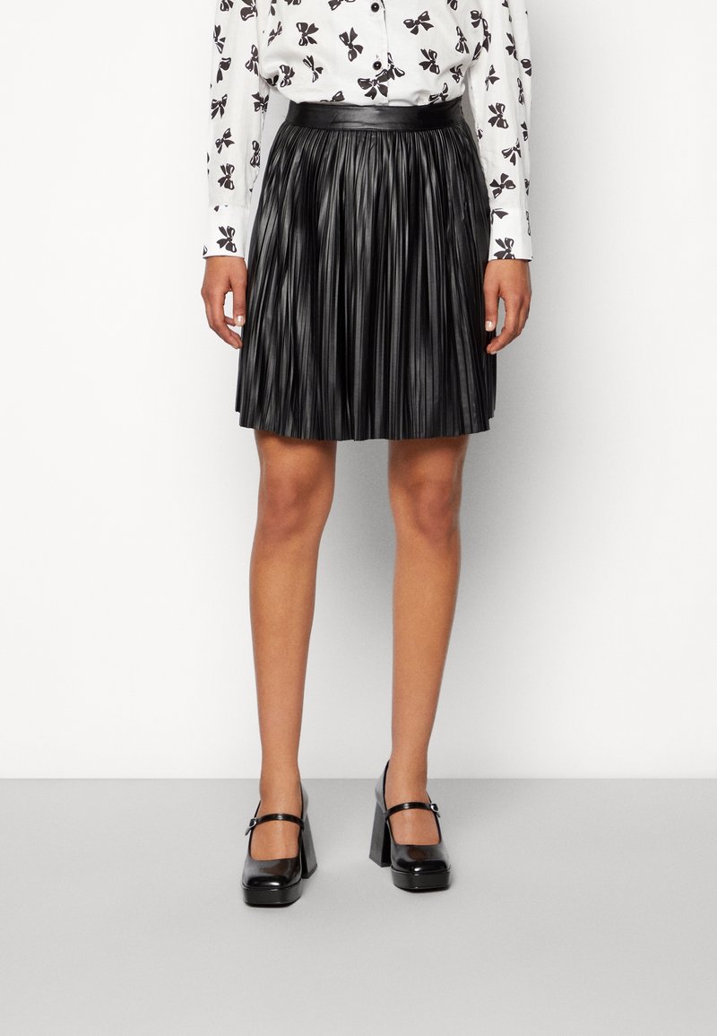 Femme portant une jupe plissée noire, des chaussures Mary Jane noires à talons hauts, et un chemisier blanc avec un motif papillon noir.