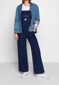 Granatowe ogrodniczki o szerokich nogawkach, z akcentem w postaci kieszeni zapinanej na zamek z przodu i naszywki z logo, w zestawie z jasnoniebieską kurtką dżinsową oraz białymi trampkami.