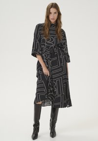 Abito nero con motivi geometrici astratti color crema, maniche corte, vita con cintura e lunghezza media, abbinato a stivali neri al ginocchio.